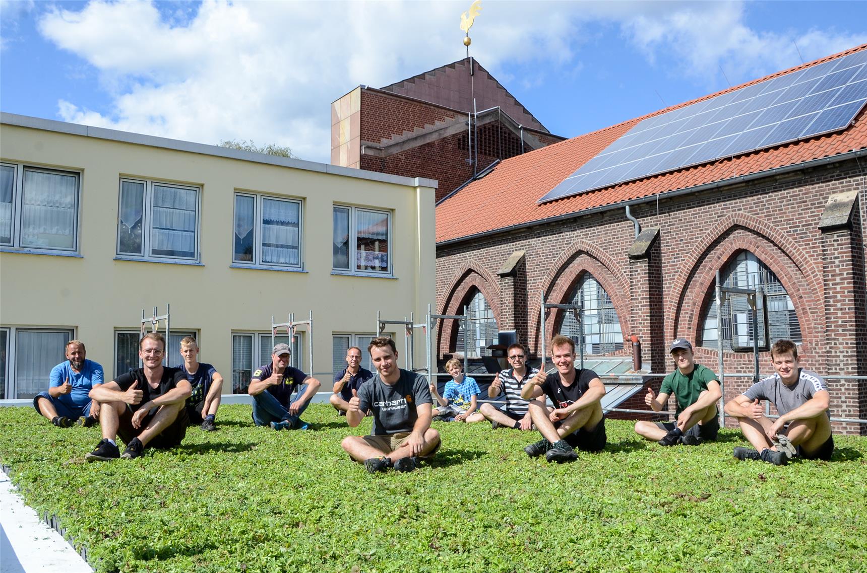 Personen sitzen auf einem begrünten Flachdach und strecken den Daumen der rechten Hand in die Höhe