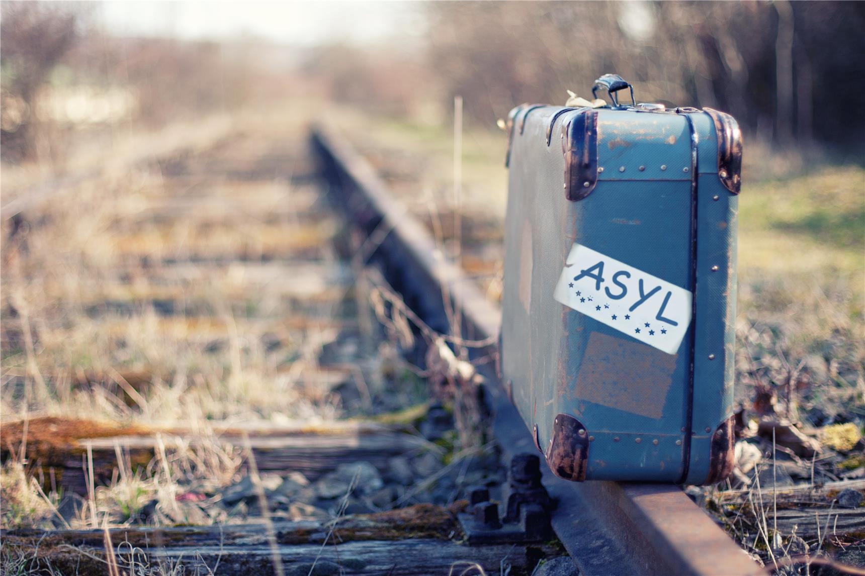 Koffer mit Aufdruck Asyl steht auf Eisenbahnschienen.