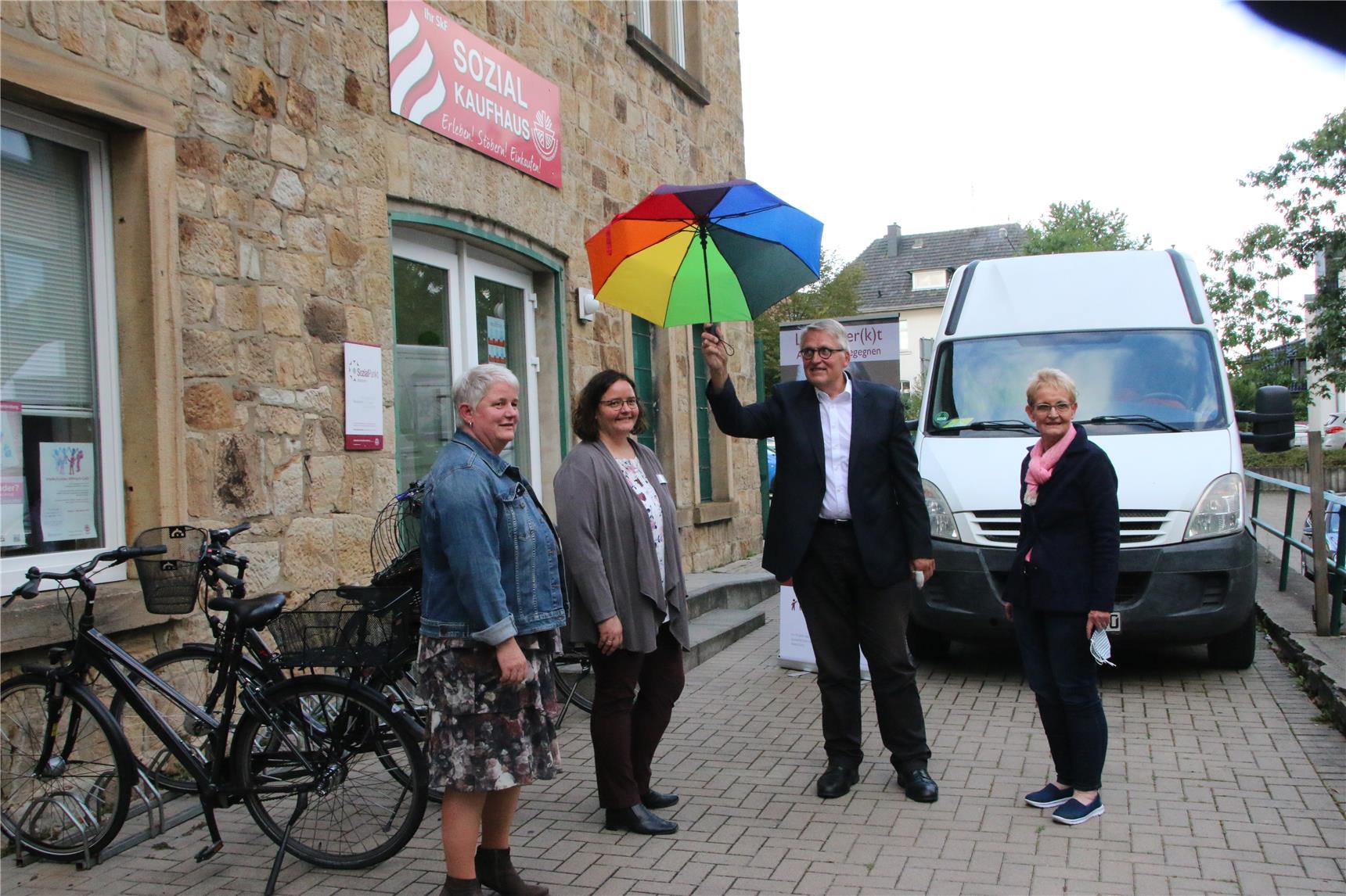 Das Foto zeigt eine Gruppe von Menschen vor einem Gebäude, einer hält einen bunten Schirm in der Hand.