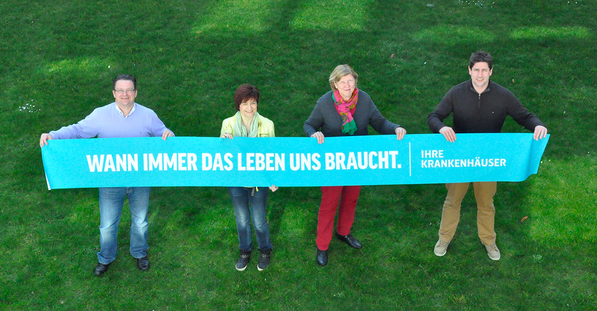 Zwei Männer und zwei Frauen halten auf einer Wiese ein Banner der Kampagne der NRW-Krankenhäuser mit dem Namen Wann immer das Leben uns braucht hoch