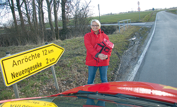 Sandra Rath steht vor ihrem roten Dienstwagen an einer Landstraße. Neben ihr ist ein Schild mit zwei Orts- und Kilometerangaben zu sehen, im Hintergrund erstreckt sich eine Wiesenlandschaft.