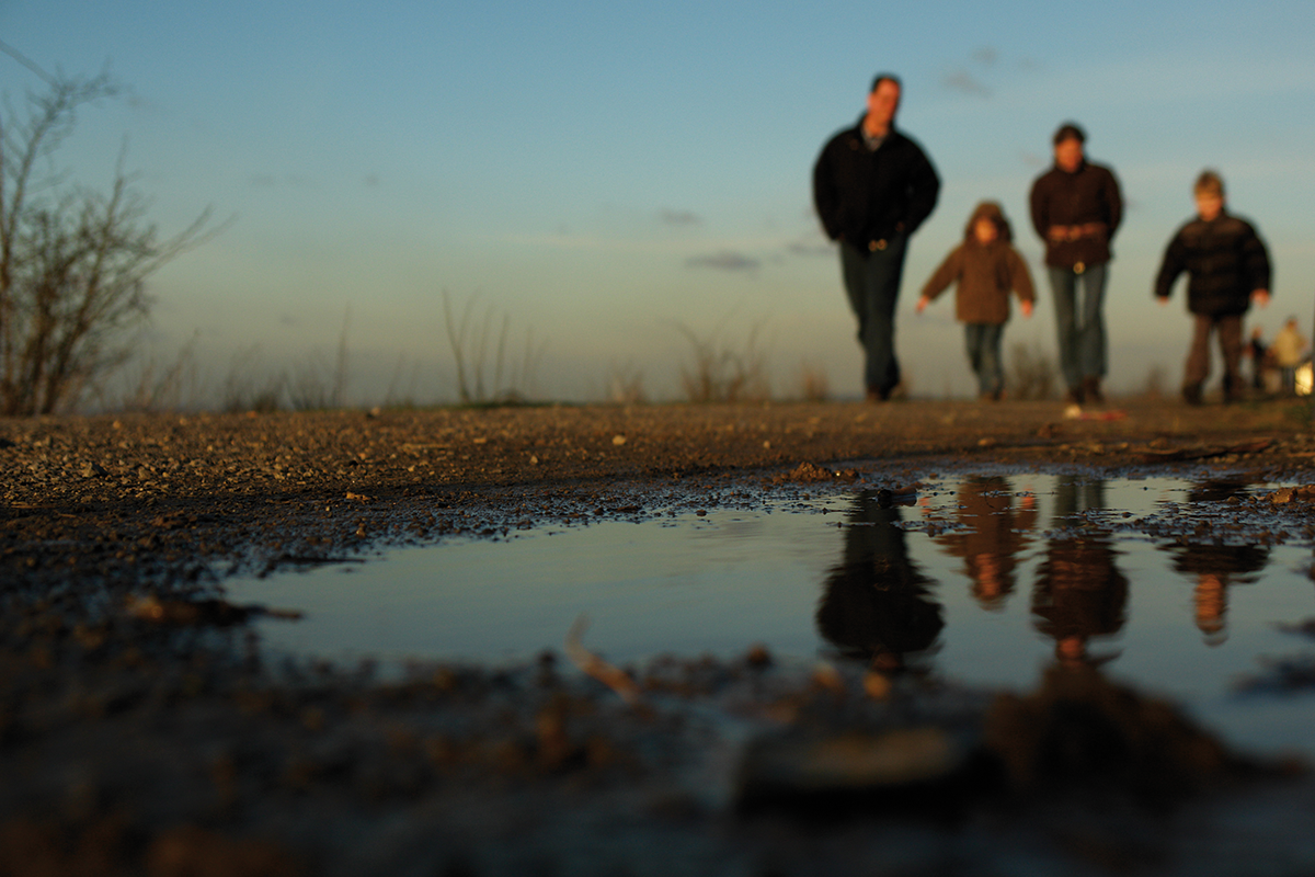 Eine Familie bestehend aus zwei Erwachsenen und zwei Kindern gehen einen Weg entlang. Im Vordergrund ist eine Pfütze zu sehen. Das Foto wurde schräg von unten aufgenommen.