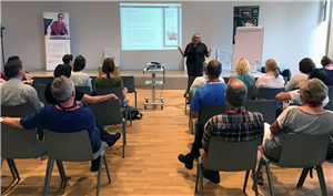 Sabine Depew steht neben einem Rollwagen, auf dem ein Beamer steht, und hält eine Präsentation auf dem Barcamp Soziale Arbeit 2017 in Siegburg. Im Vordergrund ist das Publikum zu sehen.