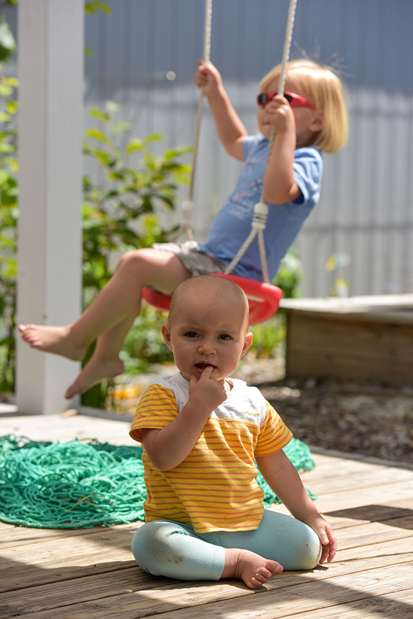 Ein kleiner Junge sitzt auf dem Boden einer Holzterasse und blickt in die Kamera. Im Hintergrund sitzt ein kleines Mädchen auf einer rot-weißen Schaukel.