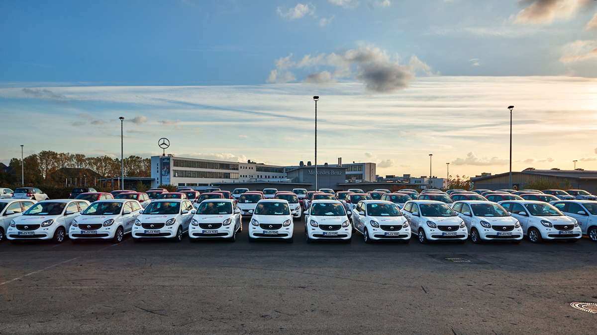 Eine große Zahl an Elektro-Smarts, die aufgereiht auf einem Parkplatz stehen. Im Hintergrund ist ein Firmengebäude von Mercedes-Benz zu sehen.