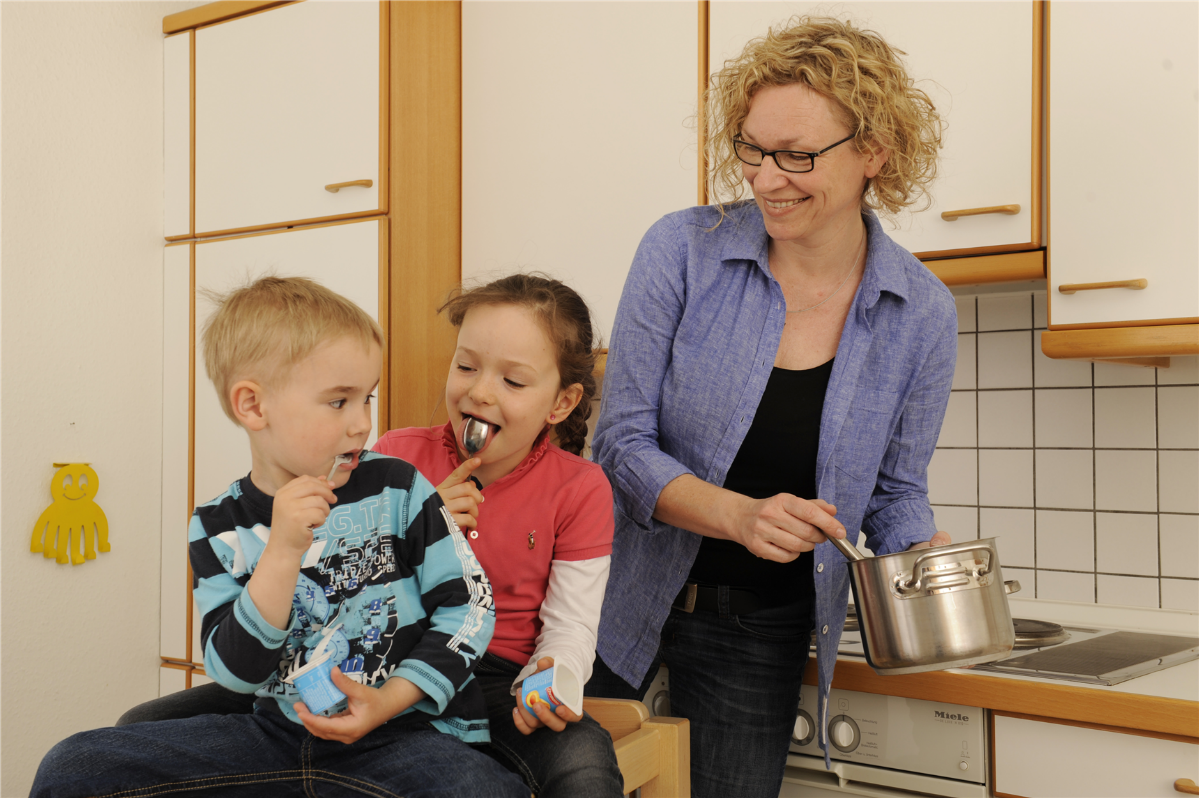 Eine Frau steht mit einem Kochtopf in einer Küche. Daneben sitzen ein Junge und ein Mädchen, die beide jeweils einen Joghurt essen.