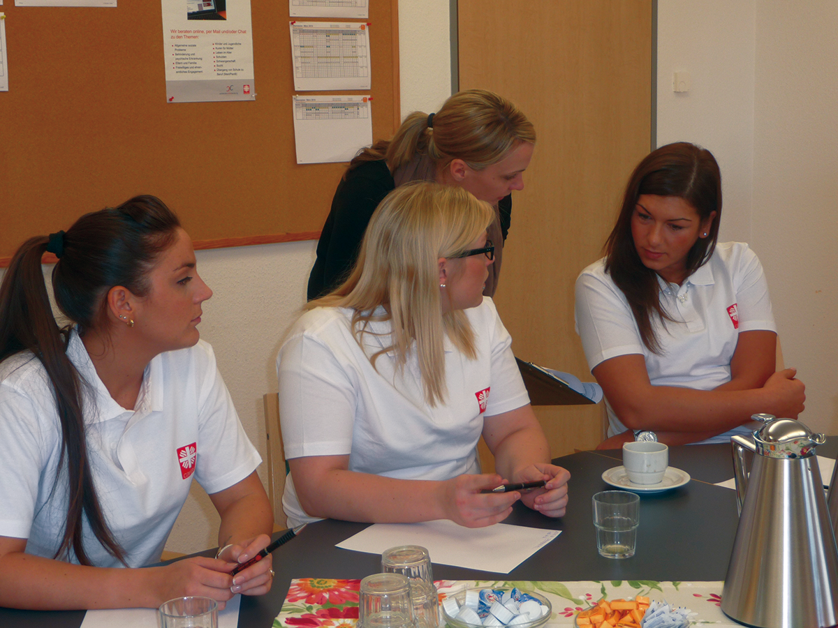 Drei Pflegerinnen der Caritas sitzen an einem gedeckten Besprechungstisch. Dahinter steht eine Frau mit Unterlagen in der Hand, die Sie mit den Pflegerinnen bespricht.