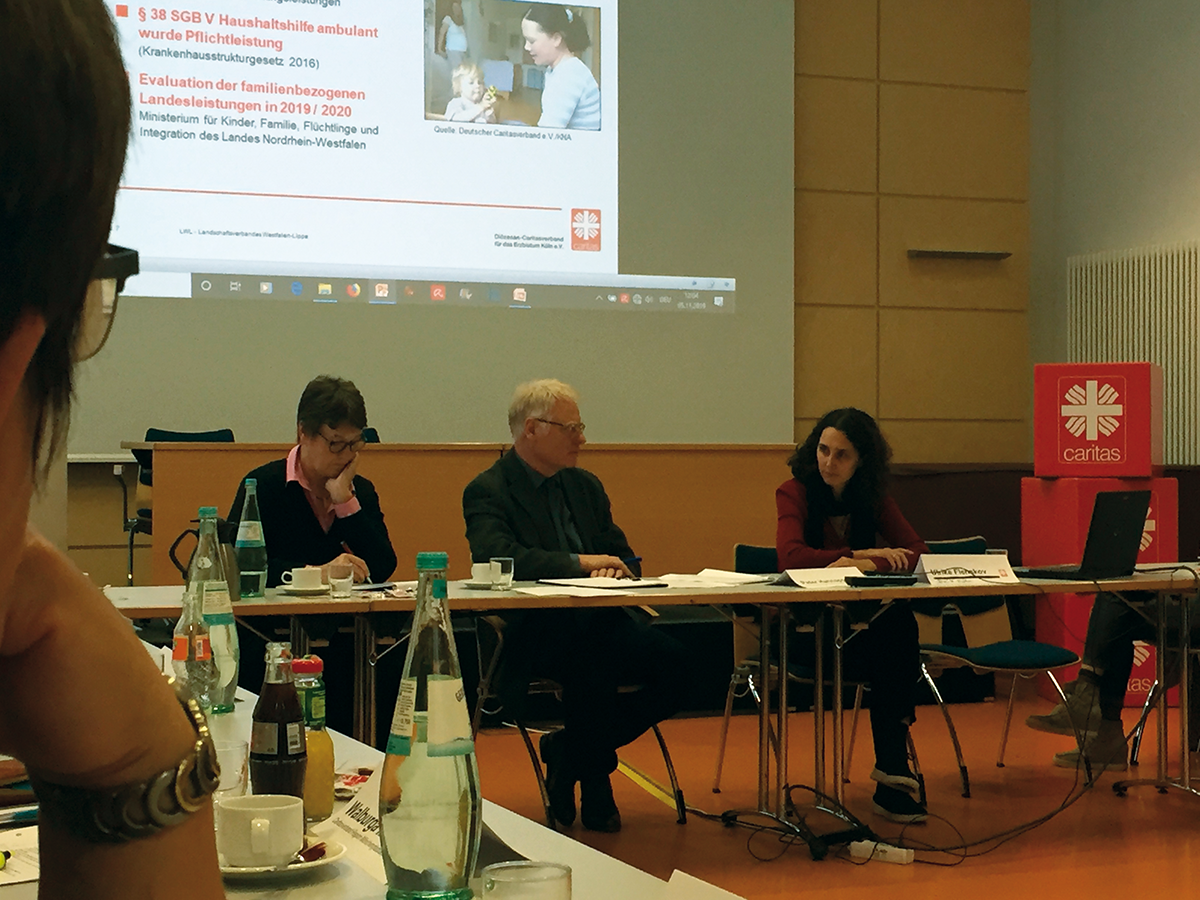 Zwei Referentinnen und ein Referent sitzen bei der Konferenz der Familienpflegedienste der CiNW an einer Tischreihe. Im Hintergrund ist die Bühne und die Projektion eines Beamers zu sehen.