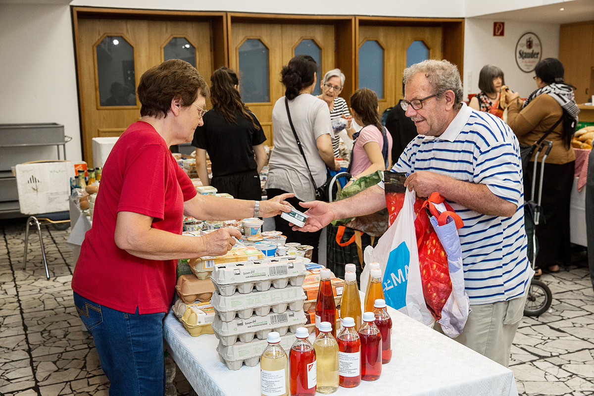 Ein Senior nimmt in einem Tafelladen gekaufte Waren von einer Ehrenamtlichen entgegen. Im Hintergrund sind weitere Kunden und Helfer des Tafelladens zu sehen.
