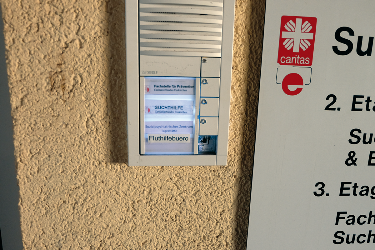 Eine Türklingel an einem Gebäude der Caritas Euskirchen, neben der ein Eingangsschild hängt. Unter den Namen der drei dort ansässigen Einrichtungen wurde die Aufschrift 'Fluthilfebüro' geklebt.