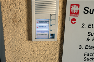 Eine Türklingel an einem Gebäude der Caritas Euskirchen, neben der ein Eingangsschild hängt. Unter den Namen der drei dort ansässigen Einrichtungen wurde die Aufschrift 'Fluthilfebüro' geklebt.