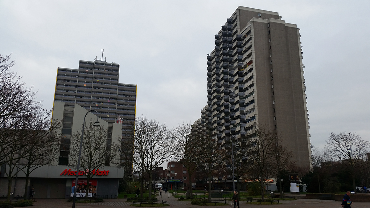 Zwei Wohnblöcke mit Plattenbauten in Köln-Chorweiler, vor dem sich eine kleine Parkanlage befindet. Auch ist die Filiale eines Elektronikhandels zu sehen.