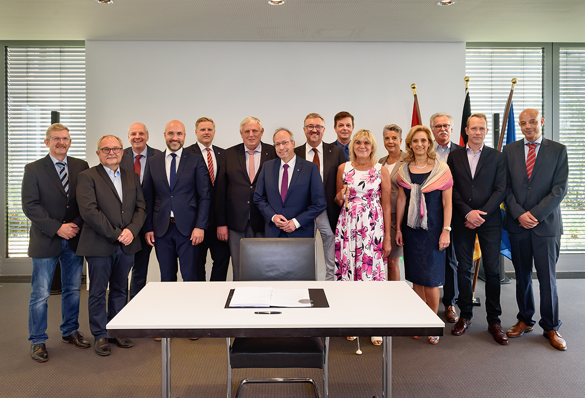 Ein Gruppenbild mit den Unterzeichner/innen des Landesrahmenvertrages. Vor der Gruppe steht ein weißer Tisch auf dem der Vertragstext liegt, davor steht ein schwarzer Lederstuhl.