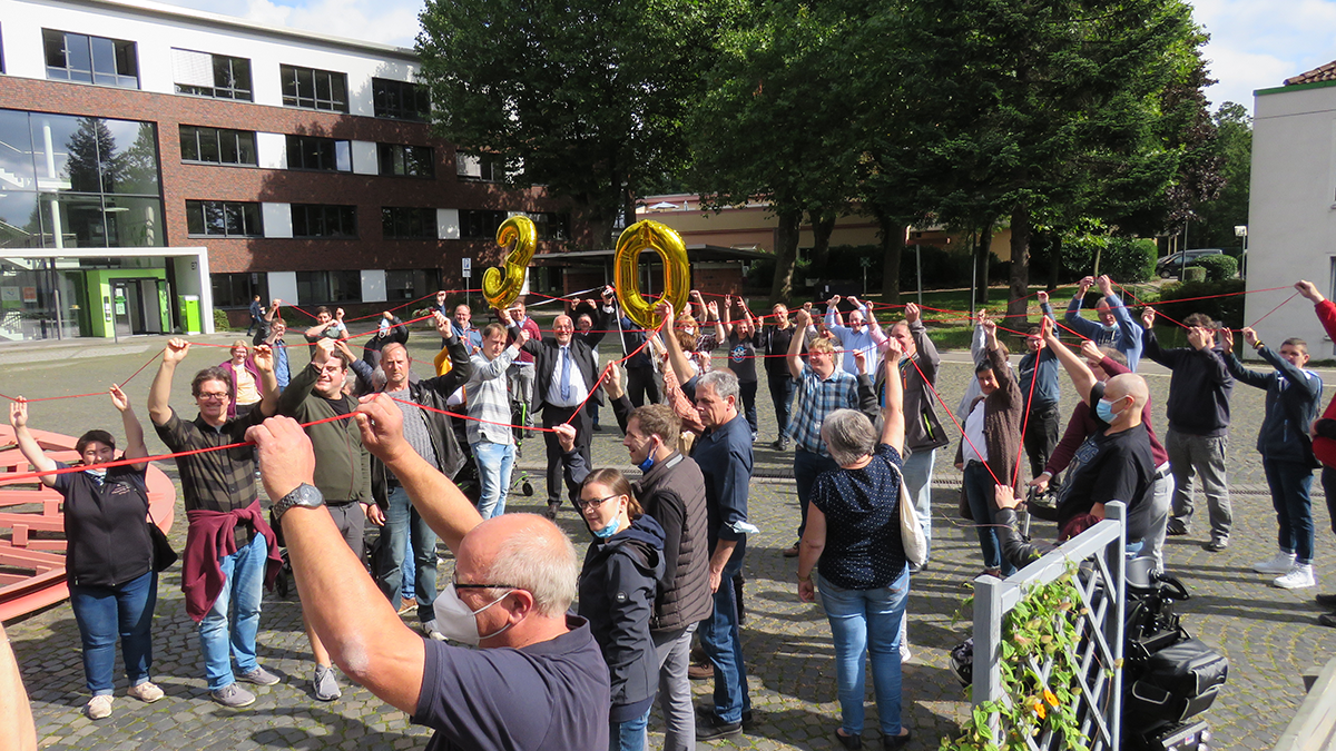 Die Teilnehmenden des 30-jährigen Jubiläums der Caritas-Werkstatträte, die auf einem Außengelände eine rote Schnur und einen goldenen Luftballon (Zahl 30) hochhalten