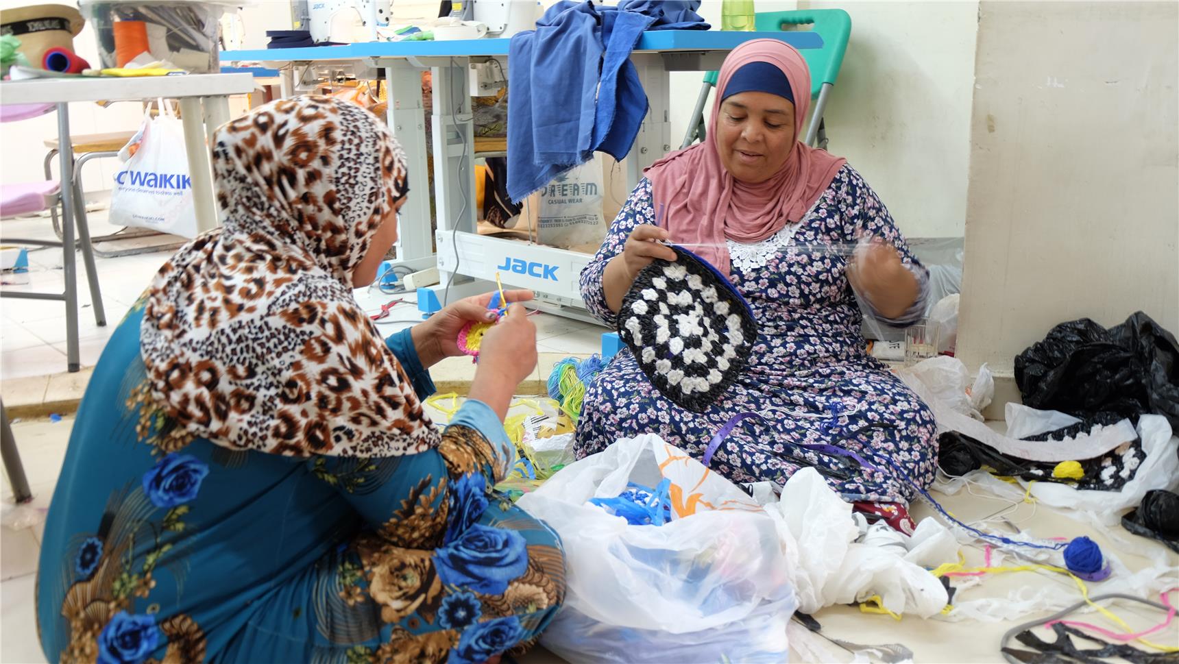 2 Frauen mit Kopftüchern sitzen auf dem Boden vor einem großen Berg Plastik und handarbeiten.