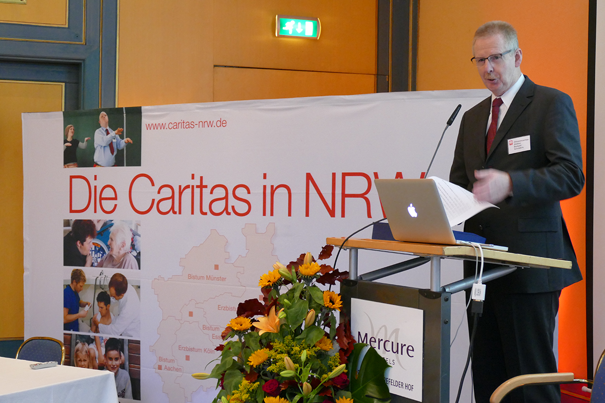 Diözesan-Caritasdirektor Burkard Schröders steht vor einem Pult mit einem Laptop, das neben einem Display der Caritas in NRW steht, und hält eine Rede