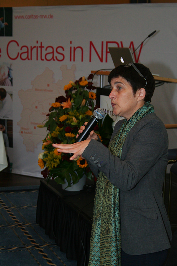 Ministerin Barbara Steffens steht mit einem Mikrofon neben einer Bühne mit Rednerpult und hält eine Rede