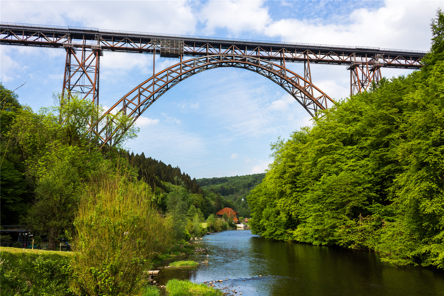 Die Müngstener Brücke (stählernde Bogen-/Eisenbahnbrücke) in Solingen, unter der die Wupper durchfließt. Die Uferseiten sind dicht bewachsen, im Hintergrund ist das Restaurant 