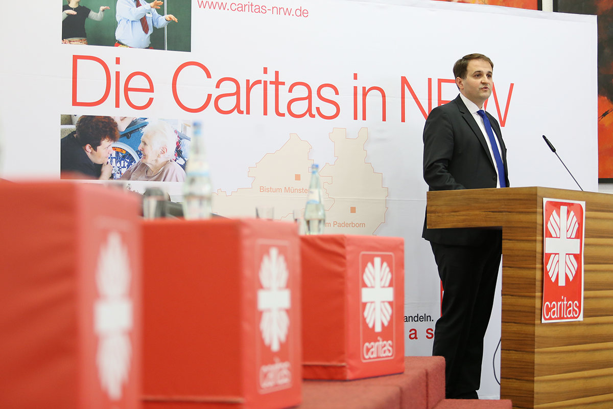 NRW-Staatssekretär Nathanael Liminski steht beim Ruhrgebietstag vor einem Rednerpult und hält sein Grußwort. Es sind außerdem ein Faltdisplay der Caritas in NRW und drei rote Caritas-Würfel zu sehen.