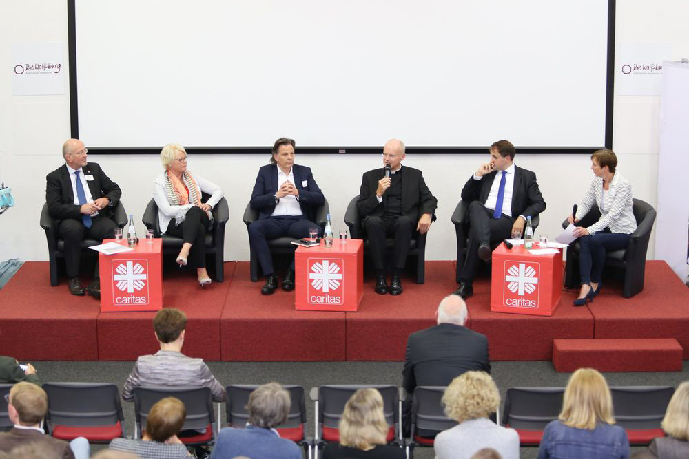 Die Teilnehmer/innen der Podiumsdiskussion des Ruhrgebietstages sitzen auf der Bühne zusammen und diskutieren. Auf der Bühne sind auch drei rote Caritas-Würfel und im Vordergrund das Publikum zu sehen