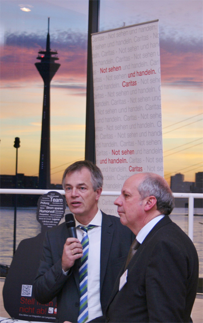 NRW-Umweltminister Johannes Remmel steht mit Diözesan-Caritasdirektor Heinz-Josef Kessmann auf einem Podium und spricht in ein Mikrofon