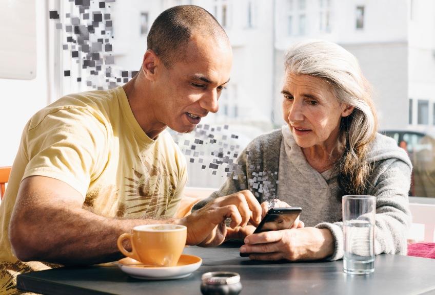 Ein junger Mann mit Migrations-Hintergrund erklärt einer Seniorin ihr Smartphone.