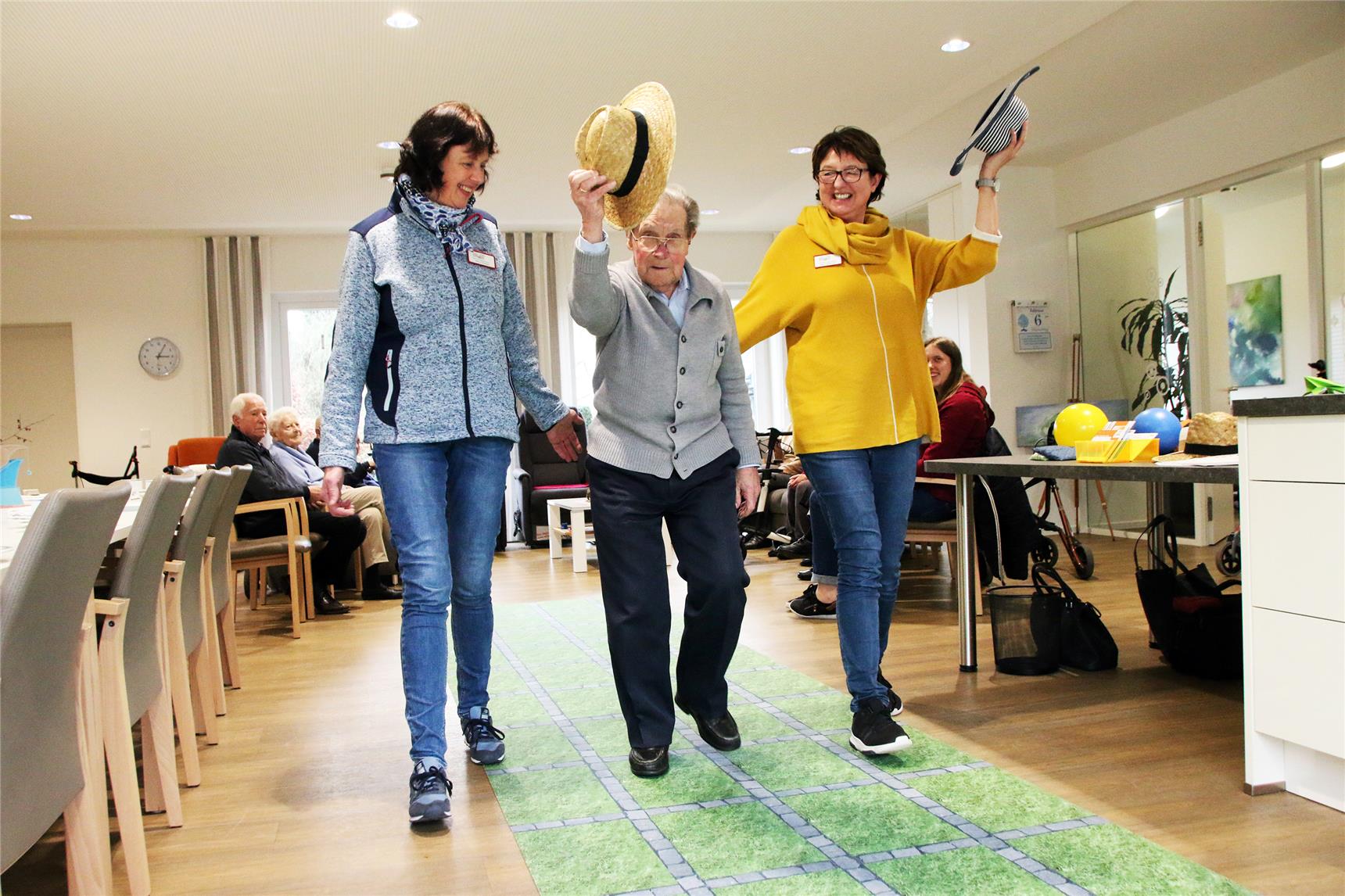 Das Foto zeigt zwei Frauen, die einen älteren Mann rechts und links begleiten, der seinen Hut lüftet.